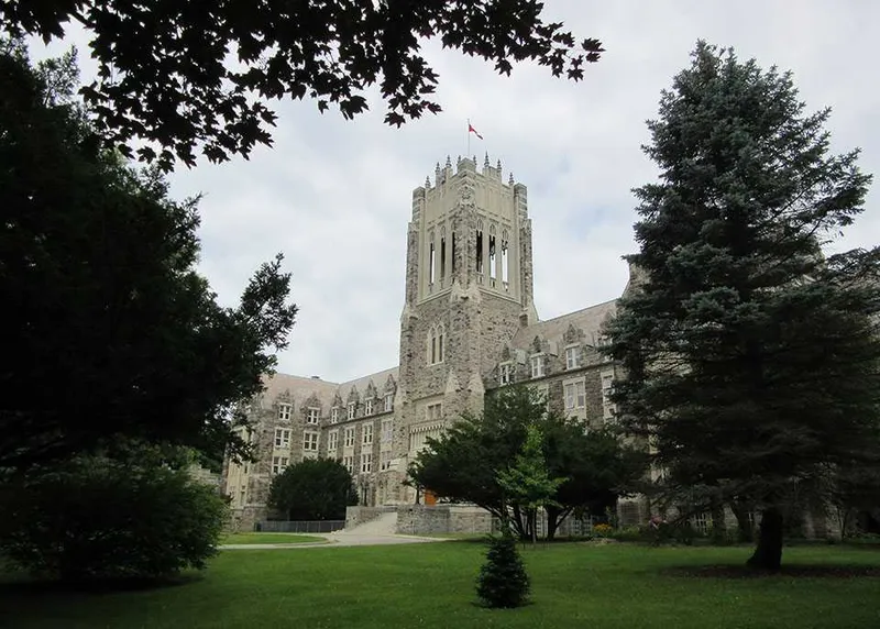 Stone Gothic-style building with a tall central tower topped by a flag, surrounded by green trees and a lawn under a cloudy sky.