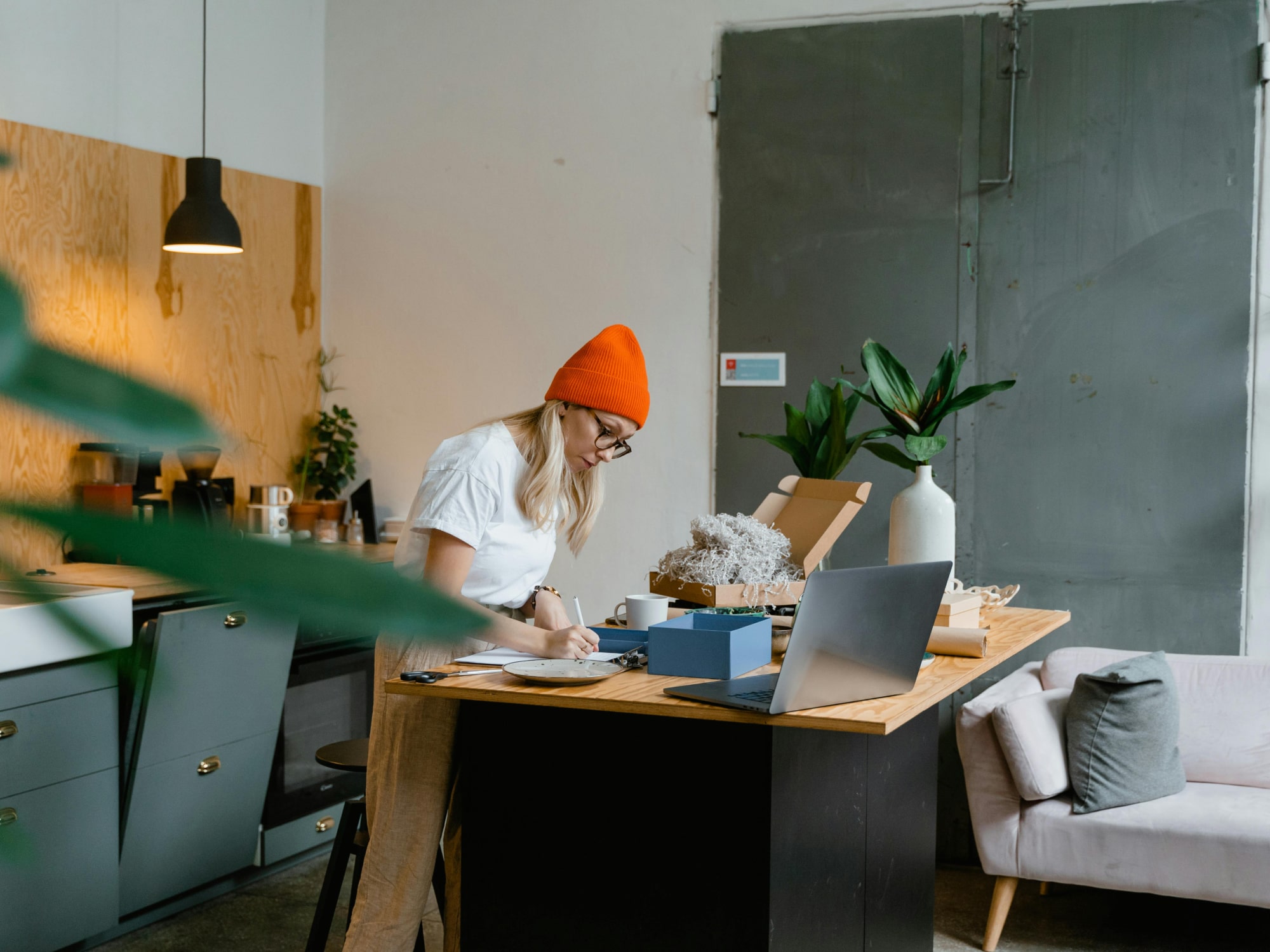 Woman in an orange beanie writing on a clipboard at a kitchen island surrounded by plants and a laptop.