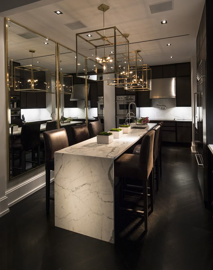 Modern kitchen with a long marble island, dark leather bar stools, brass-framed pendant lights, and large mirrors on one wall.