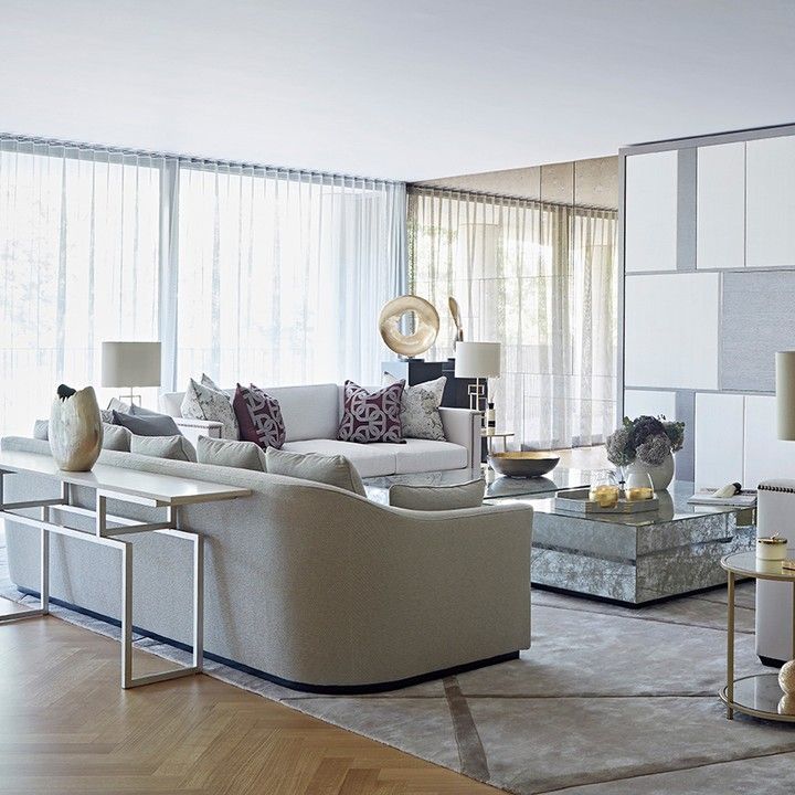Modern living room with light-colored curved sofas, patterned throw pillows, a marble coffee table, decorative vases, and sheer floor-to-ceiling curtains.