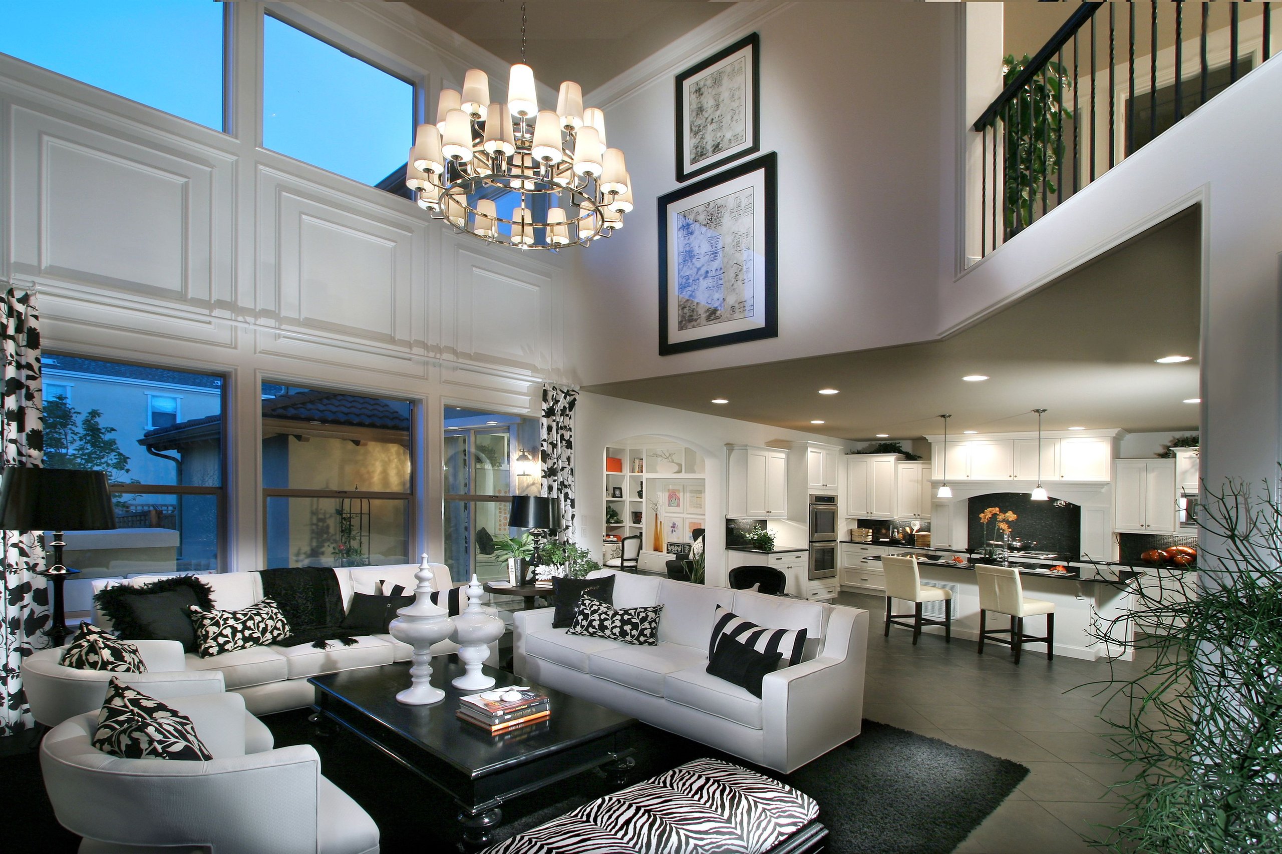 Modern open living room and kitchen with white furniture, black and patterned cushions, large windows, and chandelier lighting.