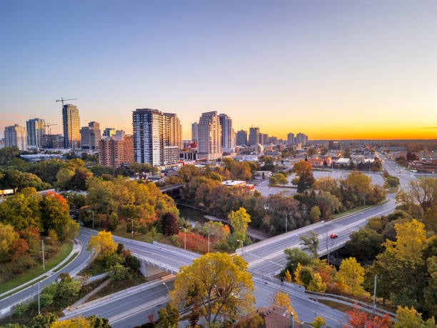 Aerial view of a cityscape at sunset with tall buildings, colorful autumn trees, and intersecting roads.