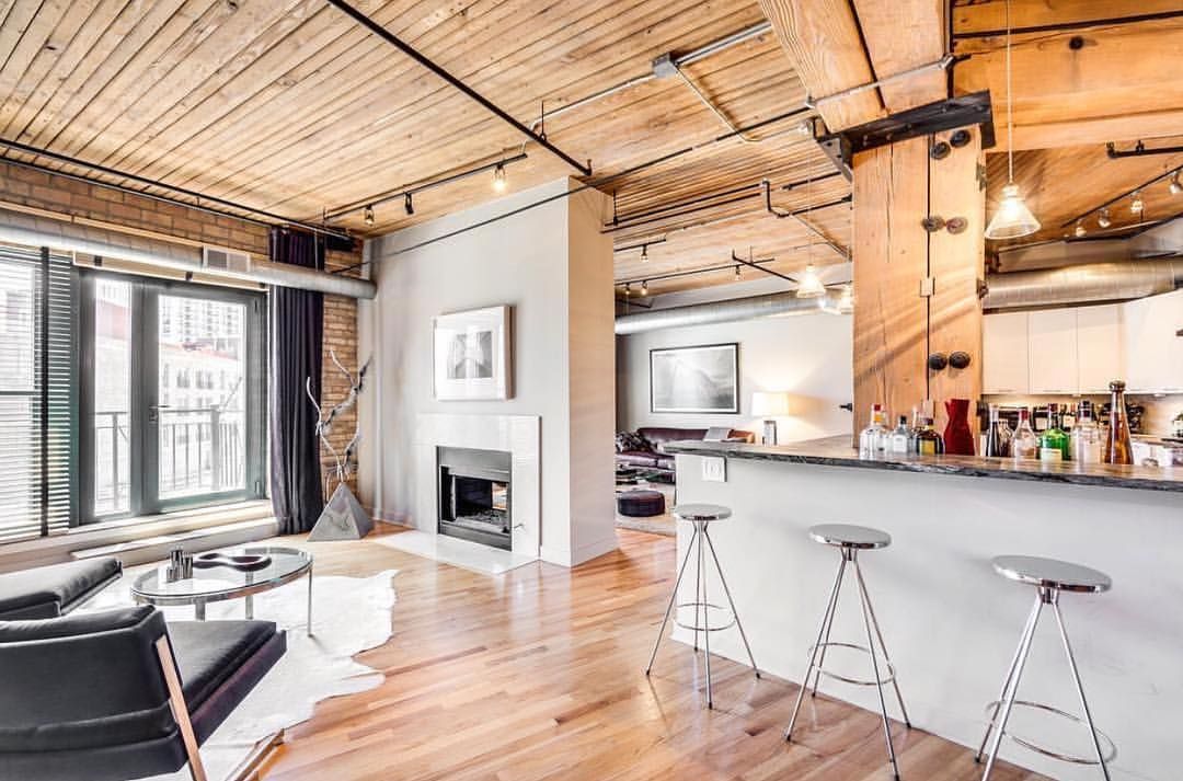Modern loft living room with wooden ceiling, fireplace, black leather chairs, glass coffee table, bar with three metal stools, and kitchen in the background.