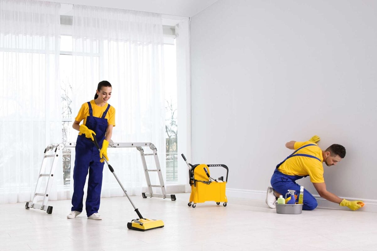 Two cleaners in yellow shirts and blue overalls cleaning a bright room; one sweeping the floor and the other scrubbing the wall baseboard.