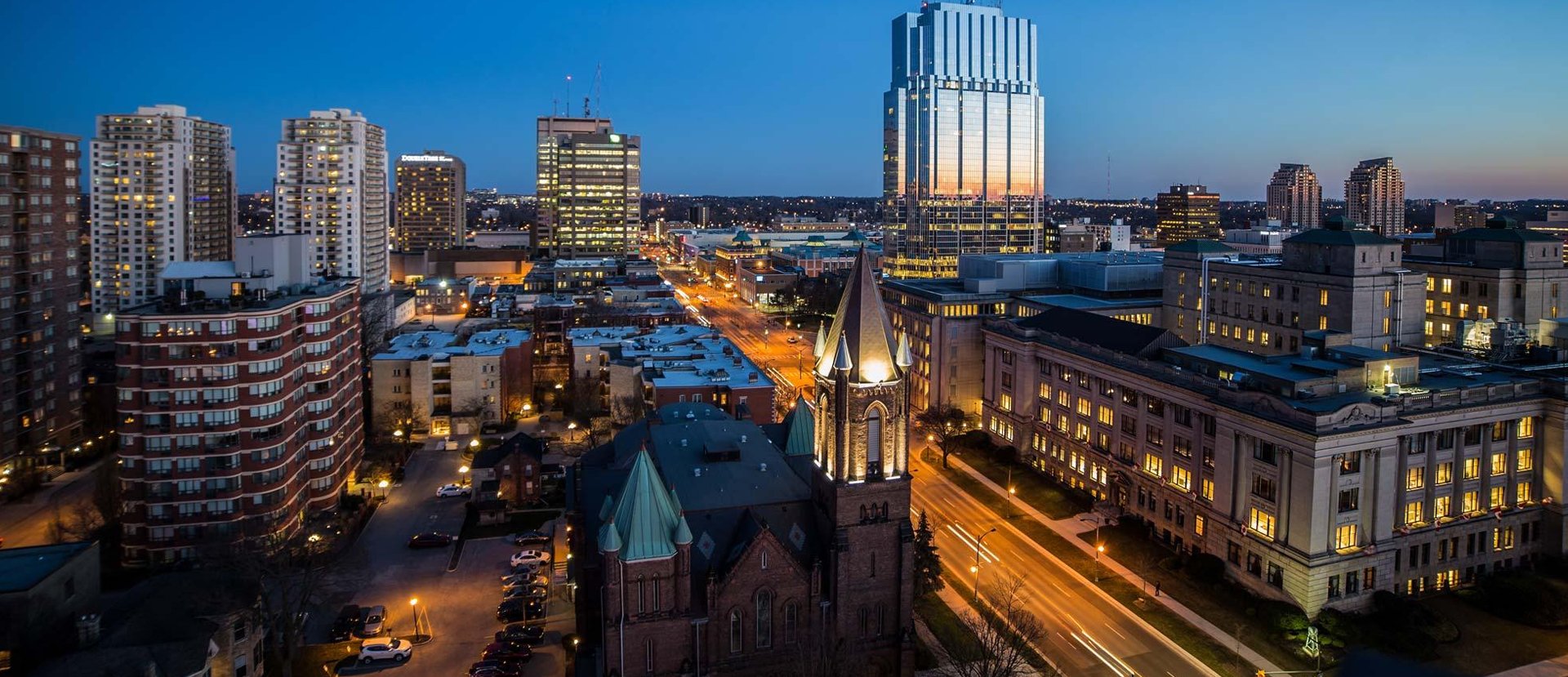 Cityscape at dusk with illuminated church tower and tall glass skyscraper reflecting sunset colors.
