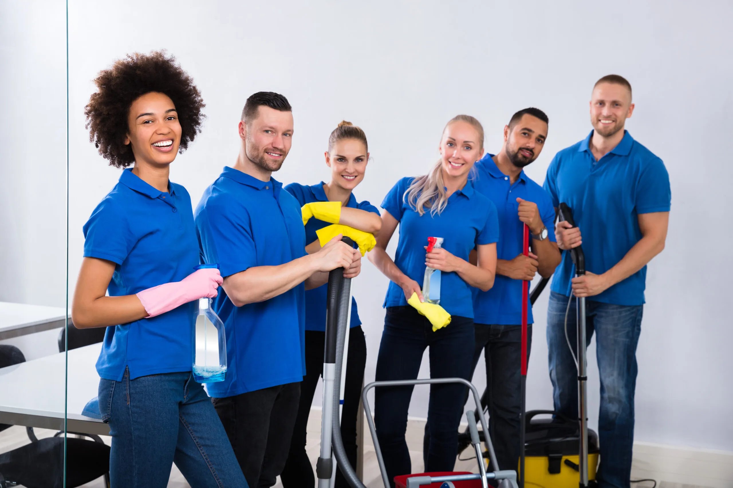 Six diverse professional cleaners in blue shirts holding cleaning tools and supplies, smiling indoors.