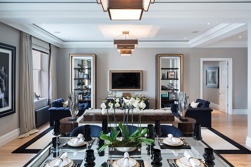 Modern living and dining room with glass table set for six, white orchid centerpiece, two navy armchairs, and a wall-mounted TV flanked by mirrored shelves.