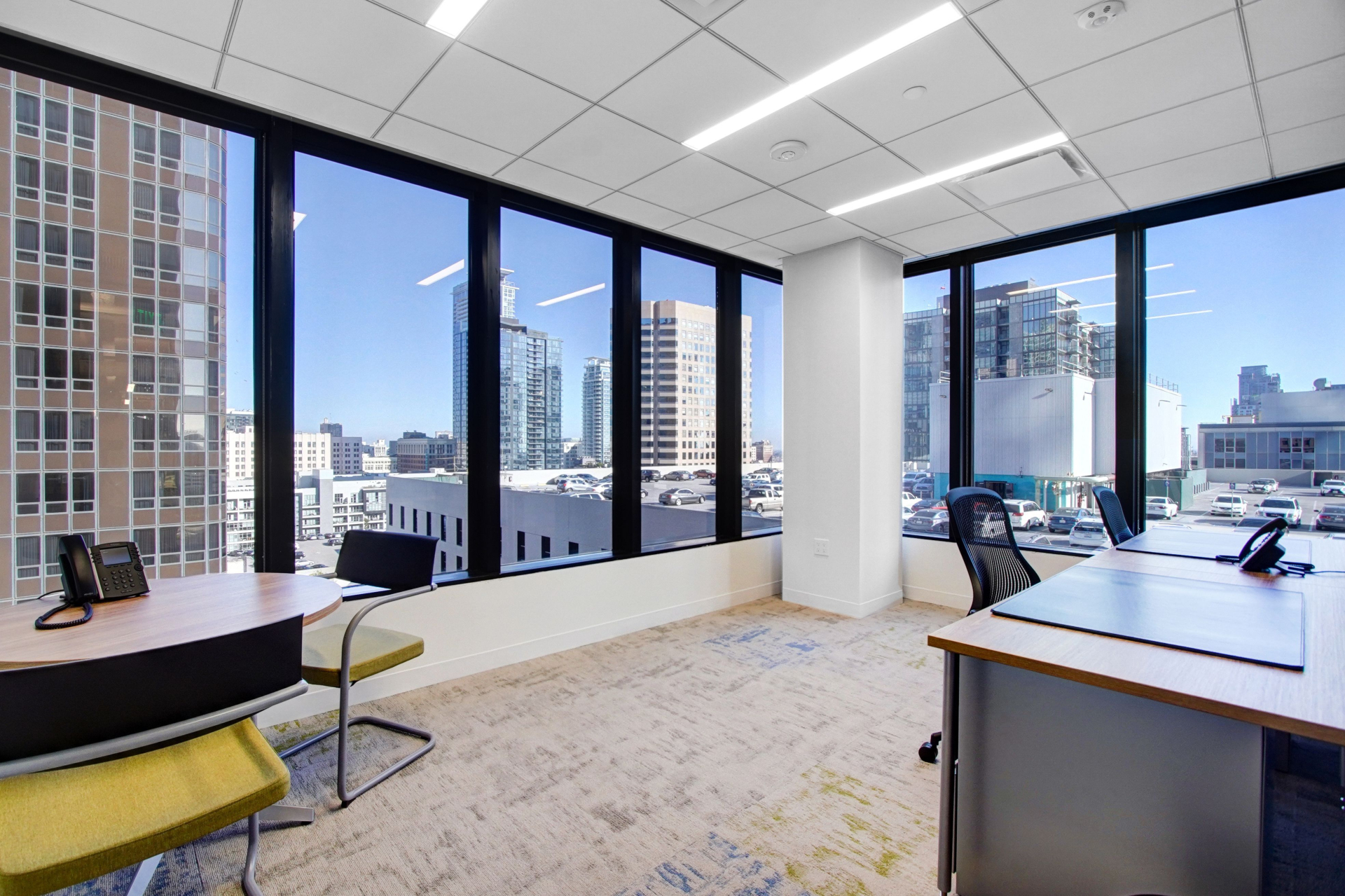 Modern corner office with large windows overlooking city buildings and parked cars, featuring desks, chairs, and office phones.
